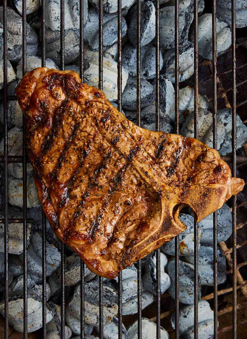 Juicy, perfectly grilled T-bone steak cooking over hot charcoal on a barbecue grill served at Mahony’s Steak House Bishopbriggs